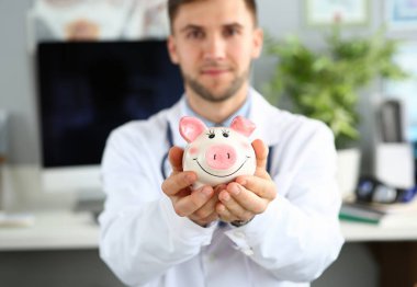 Kumbara tutan doktor ellerine odaklan. Neşeli doktor kar beyazı üniformayla hastane ofisinde poz. Sağlık sigortası poliçesi ve tıp kavramı. Bulanık arka plan