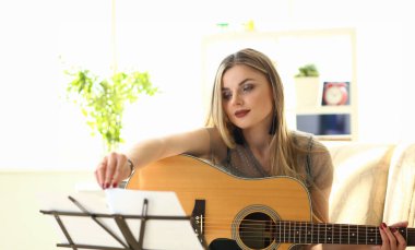 Kadın Uygulama Akustik Gitar Evde Çalma. Oturma Odasında Kanepede Otururken Performans Yapan Kadın Oyuncu. Sarışın Lady Müzik Standı'nda Notlar Dönüyor. Kafkas Kız Akorları Alarak