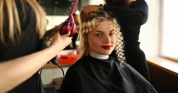 Young Woman Getting Curly Hair by Curling Iron. Styling Wavy Hairstyle for Beautiful Client. Girl Sitting with Funny Long Lock. Hairstylist Making Hairdo in Beauty Salon Looking at Camera Shot