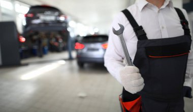 Beyaz koruyucu eldivenli servisçi, araba servis istasyonu arka planına karşı elinde matall anahtarını tutar
