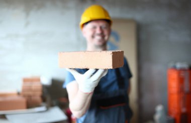 Beton bloğu sunan erkek el odaklanın. Bir elinde kırmızı tuğla tutan ve memnuniyetle kameraya bakarak koruyucu kıyafetli mutlu gülümseyen yapıcı adam. Bina konsepti