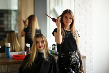 Genç Kızlar Kuaför ve Güzellik Salonu Müşteri. Kuaför Elinde Kıvırma Demir tutan. Kadın Müşteri Saç Kesimi Yapmak Için Stüdyoda Oturuyor. İki güzel sarışın kadın. Şekillendirme Saç Modeli