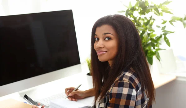 Black smiling woman sitting at workplace writing something in diary portrait
