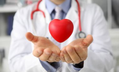 Kırmızı kalp şeklini koruyan erkek doktor ellerinin yakın çekimi. Beyaz üniforma ve steteskopla poz veren bir tıp çalışanı. Tıp kardiyolojisi ve sağlık konsepti
