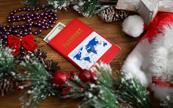 Close-up of red passports cover with banknotes in it. White and blue plastic credit card on top. Bright festive christmas decorations on wooden table. Holiday concept
