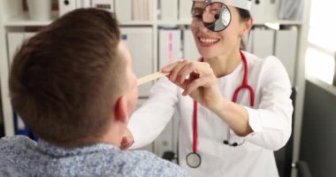 Ön aynalı doktor, hastanın boğazını ahşap spatula 4K filmle yavaş çekimde inceliyor. Yetişkinlerde kronik bademcik iltihabı teşhisi ve tedavisi