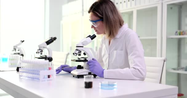 科学研究室の助手科学者医師が調査の結果を書いている 女性は顕微鏡を見て医学的分析を調べる — ストック動画