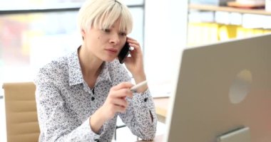 İş kadını ofis 4K filminde cep telefonuyla bilgisayarın önünde ağır çekimde konuşuyor. İş görüşmesi ve planlama çağrısı