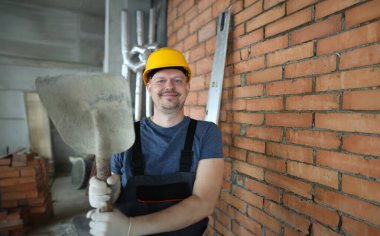 Sert şapkalı işçi çimento küreği tutuyor. İnşaat alanında gülümseyen erkek müteahhit