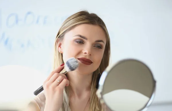 Smiling young woman looks in the mirror and paints herself with blush brushes. Makeup artist girl doing makeup learning with beauty and cosmetics video tutorials