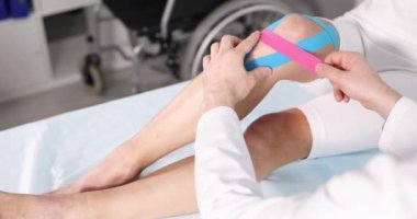 Doktor klinikte yaralı dizine Kinesiotape yapıştırıyor. 4K film yavaş çekimde. Travma konseptinden sonra Kinesiyoterapi