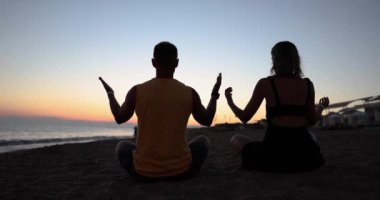 Young couple doing yoga on seashore at sunset back view 4k movie slow motion. Calmness and meditation concept