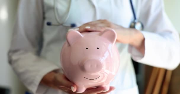 医者はピンクの貯金箱をコインで撫でて閉じる4K映画スローモーション 有料医療の概念 — ストック動画