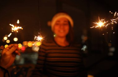 Eldeki maytap parlak bir şekilde yanıyor. Arka planda Noel Baba şapkalı bir kadın var..