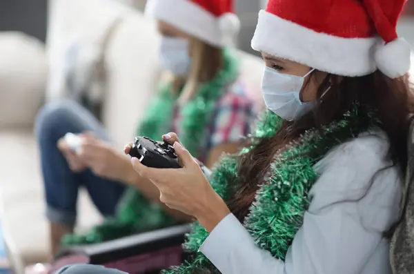 Two girlfriends play game console. Woman in protective mask and santa claus hat hold joystick.