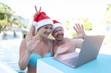 Noel Baba şapkalı bir adam ve kadın tatilde dizüstü bilgisayara el sallıyorlar. Yeni yıl tatili uzaktan tebrik konsepti