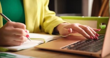 Sarı takım elbiseli bir kadın bir deftere yazı yazıyor ve dizüstü bilgisayara yakın çekim yapıyor. Ofis işi, iş eğitimi.