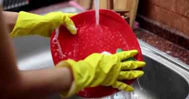 Temizlikçi kadın akan suyun altında kırmızı tabağı yıkıyor. Süngerle yakın çekim 4K film yavaş çekim. Ev temizliği bulaşık konsepti