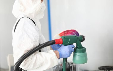 Duvarı sprey silahla boyayan profesyonel işçilere yakın plan. Beyaz koruyucu kostüm ve mavi eldiven giymiş biri. Yenileme ve inşaat alanı kavramı