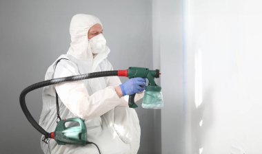 Profesyonel erkek işçinin portresi duvarları gri renkte sprey silahla boyuyor. Beyaz koruyucu kostümlü ve yüz maskeli bir yetişkin. Sağ tarafta uzayı kopyala. Yenileme konsepti