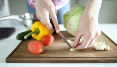 Kadınlara yakın çekim, tahta tahtada keskin gümüş bıçakla sarımsak kesme. Masada tam sarı biberli lahana salatalığı ve yumuşak domates var. Lezzetli kahvaltı konsepti hazırlanıyor