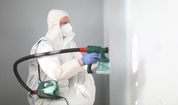 Portrait of professional male worker painting wall in grey colour with spray gun. Adult in white protective costume and face mask. Copy space in right side. Renovation concept