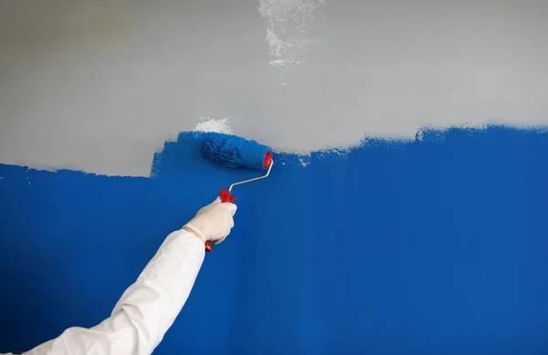 Close-up of adult painting wall with bright blue paint. Man using roller for renovation work. Change interior design in new modern apartment. Construction site concept