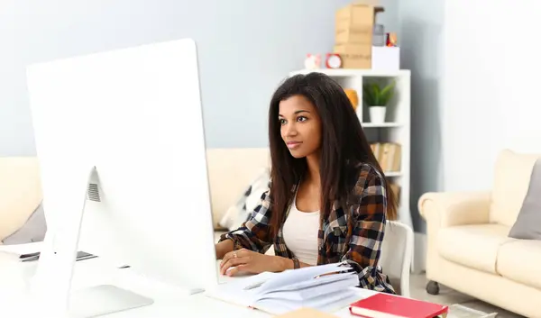 Black smiling woman sitting at workplace working with desktop pc studying or doing school homework concept