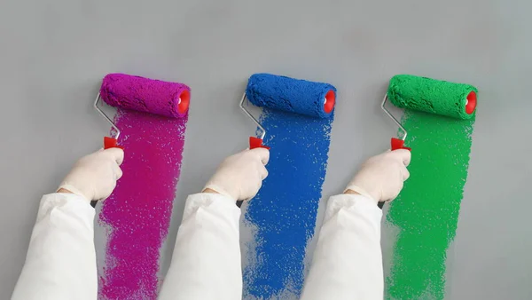Close-up of professional builder painting wall in blue, red and green paint. Person holding roller for work. Grey wall. Change interior design in apartment. Renovation and construction site concept