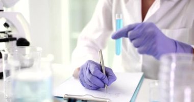 Laboratuvar asistanı notlar alır ve test tüpünü mavi sıvıyla tutar. Gözlemleri ve sonuçları yazan bilim adamı