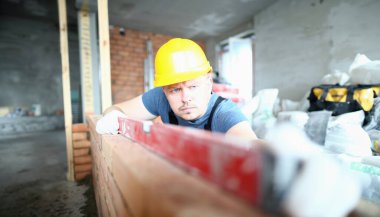 Elleri zarar görmeden tutmak için sert eldivenler giyen sakallı inşaatçıportresi. Profesyonel adam denge ve tuğla duvar yüksekliği ölçmek için özel ekipman kullanarak. Bina konsepti