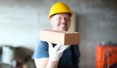Erkek elinde ciddi tuğla odaklanın. Büyük bir gülümseme ile kameraya kırmızı blok sunan hardhat işçi. Sarı kasklı neşeli yapıcı, yüzünde mutlulukla bakıyor. Bina konsepti