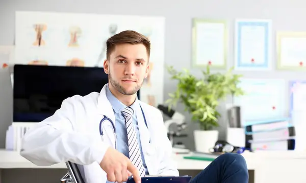 Portrait of handsome man looking in camera with joy and sitting in chair. Trendy man wearing white laboratory coat and stethoscope. Medical and healthcare treatment concept