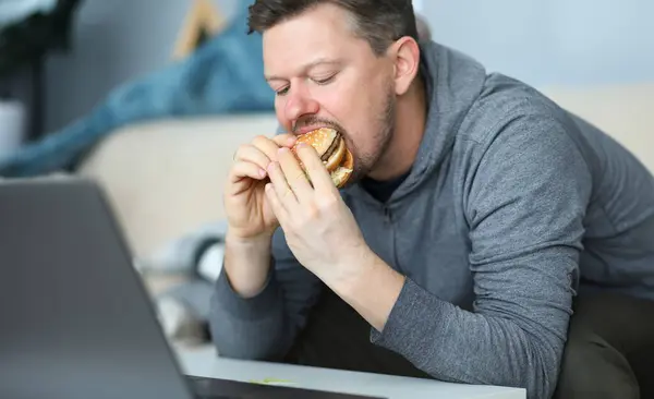 Man sit on sofa and eating burger against flat background. Unhealthy calories food concept