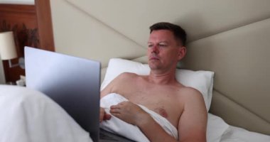 Yatak odasında ya da otelde dizüstü bilgisayar kullanarak yatağa odaklanmış bir adam. Uzaktan bağımsız çalışma ve önemli haberleri okumak
