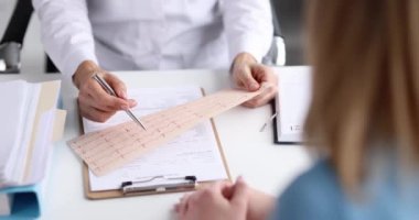 Doktor kardiyolog, 4K filminde ağır çekimde hastanın önünde EKG bandını inceliyor. Ritim ve iletkenlik bozuklukları teşhisi
