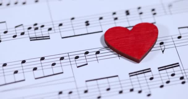 ミュージカルノート4K映画のスローモーションで紙の上に横たわる赤いおもちゃの心臓 好きな音楽コンセプト — ストック動画