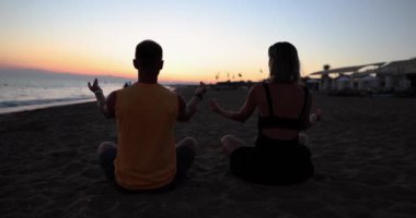 Couple yoga for meditation and meditating at serene sunset on beach. Girl and man are relaxing in lotus position in calm zen moment