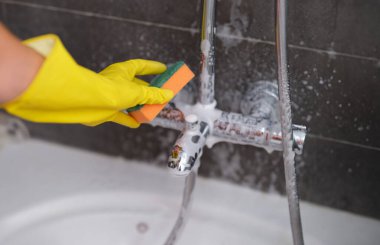 Woman in yellow glove cleans the chrome faucet in bathroom with sponge. Cleaning plumbing concept