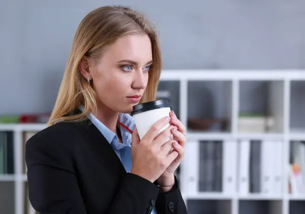 Smiling business woman drinking coffee from a paper cup in the office portrait looking into the camera holding in her hand and resting during a break.