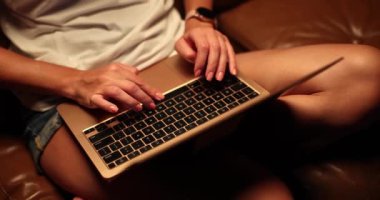 Işıklı ve dizüstü bilgisayarlı bir kadın kanepede oturuyor. Kız geceleri elleri klavye ve dokunmatik pede yakın plan dizüstü bilgisayar kullanıyor.