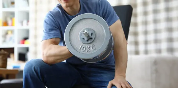 Man lifts heavy dumbbell while sitting on sofa. Interval and cardio training for burning fat. Muscle tone or weight loss. Strength training and muscle building at home. Fat burning