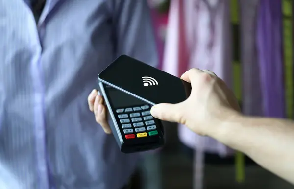 Hand holds out terminal for credit card payment. Close-up hands holding smartphone near terminal. Pay for shopping with mobile device. To pay securely for products and services by contactless payment