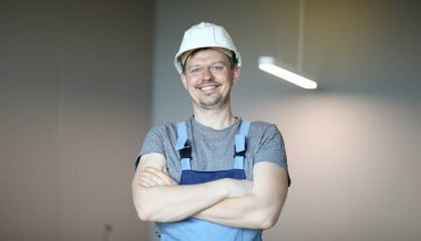 İş elbiseleri içindeki mutlu adam kollarını göğsünün üzerine kavuşturdu. İnşaatçı salgın sırasında çalışma fırsatına sevinir. İyi bir fiziksel yükleyici. Karantina sırasında kendini izole eden elektrik ve ev onarımı