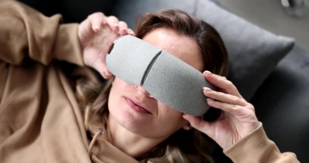 疲れた女がソファに寝そべってマッサージメガネをかける 目と顔のための多機能マッサージ — ストック動画