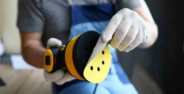 Close-up of worker holding sander machine and changing disc. Carpenter working in protective gloves. Equipment for polishing wood. Construction site and renovation concept