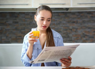 Genç ve çekici bir kız mutfakta kahvaltı sırasında gazete okuyor ve elinde bardakla portakal suyu içiyor.