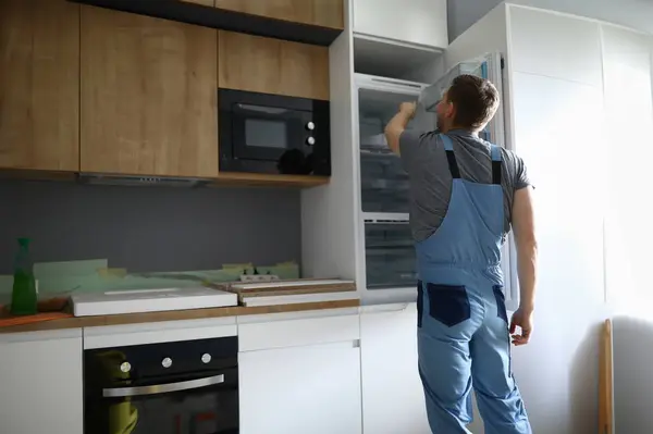 Close-up of professional handyman repair fridge. Modern kitchen set in new apartment. Worker in special uniform. Renovation moving day and disposable man concept