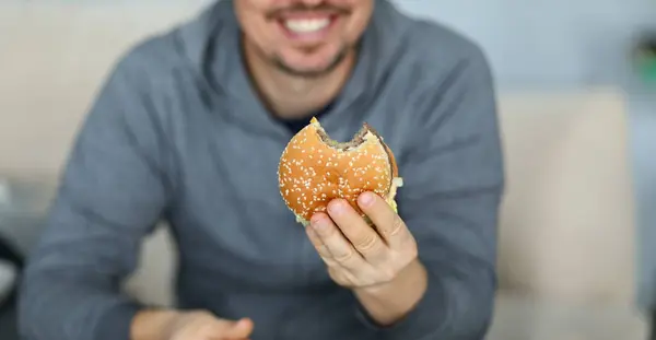 Focus on male hand holding big tasty hamburger full of mischievous calories and harmful cholesterol. Happy and fat person consuming portion of junk food