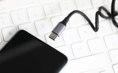Close-up view of modern mobile phone and charger plug. Smartphone with black screen laying on white keyboard of laptop. Technology and usb battery concept clipart
